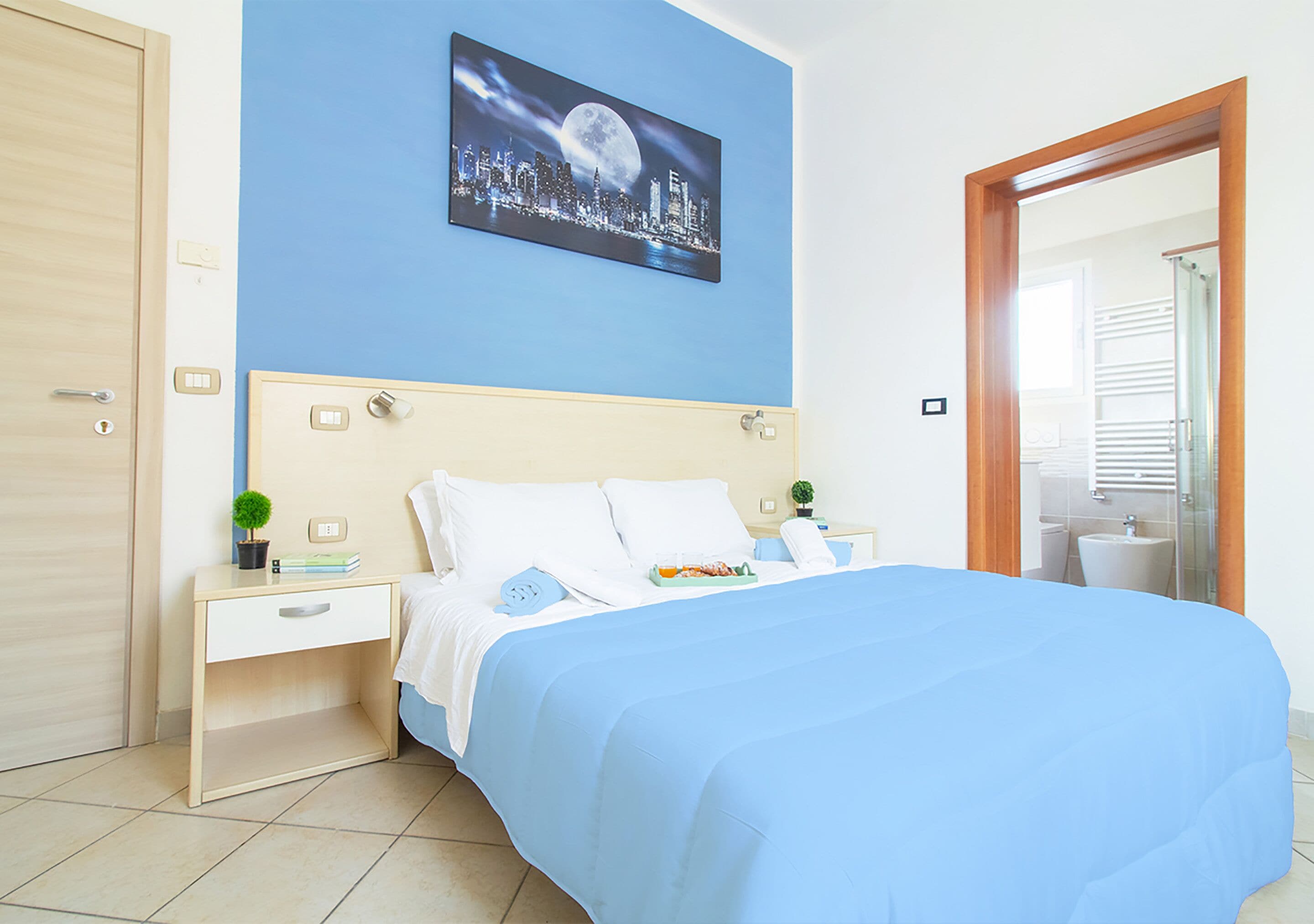 A neatly arranged bedroom with a bed, bedside tables, a nightstand, a wall-mounted cityscape picture, and a bathroom visible through an open door.