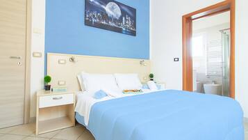 A neatly arranged bedroom with a bed, bedside tables, a nightstand, a wall-mounted cityscape picture, and a bathroom visible through an open door.