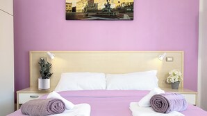 A hotel room with a bed, two rolled towels, a nightstand with a plant, a framed picture on the wall, and a small flower vase. - Athena Affittacamere (Bologna)