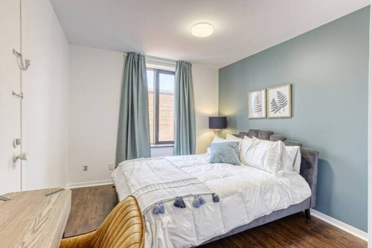 Un dormitorio con una cama, una silla, un ventanal con cortinas, una lámpara de pared y dos cuadros enmarcados.
