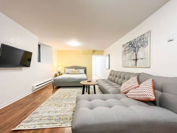 A modern living room with a grey sofa, a grey armchair, a small round table, and a bed with a grey headboard.