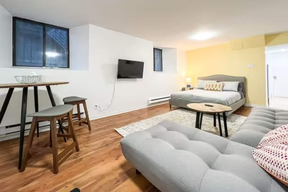 Una sala moderna con un sofá gris, taburetes de madera, una mesa de comedor y un televisor de pantalla plana montado en la pared.
