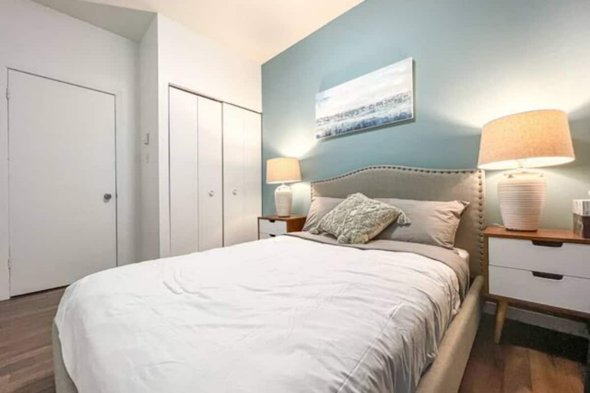Un dormitorio con una cama, dos mesitas de noche, una mesita auxiliar, una lámpara de pared y una puerta.