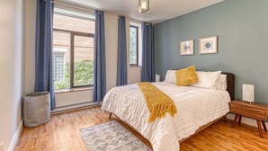 A bedroom with a large bed, a wooden floor, a window with curtains, and a small bedside table. - Le Plateau Boutique Apart Hotel (Montreal)