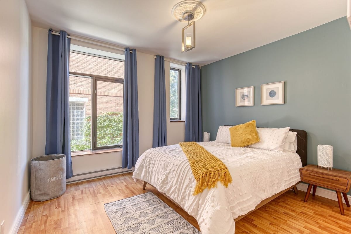 Un dormitorio con una cama grande, suelo de madera, una ventana con cortinas y una mesita de noche pequeña.