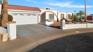 Une maison à deux étages avec un toit recouvert de tuiles rouges, un garage blanc et un jardin aménagé à l’avant, agrémenté de cactus et de palmiers.