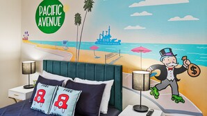 Une chambre d’hôtel comprenant un lit, des tables de chevet, une lampe et une fresque murale représentant une scène de plage avec un personnage.