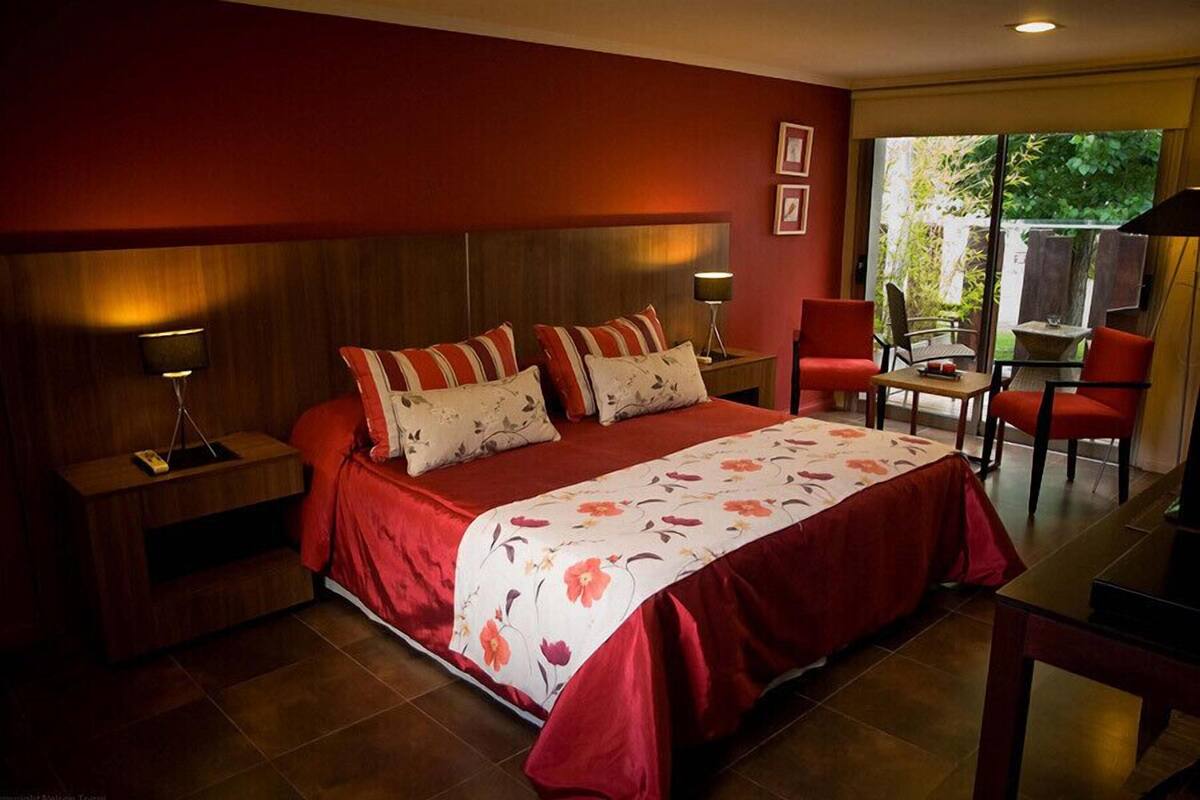 A bedroom with a large bed, a nightstand, a desk, and a chair. There is a window with a view of greenery outside.