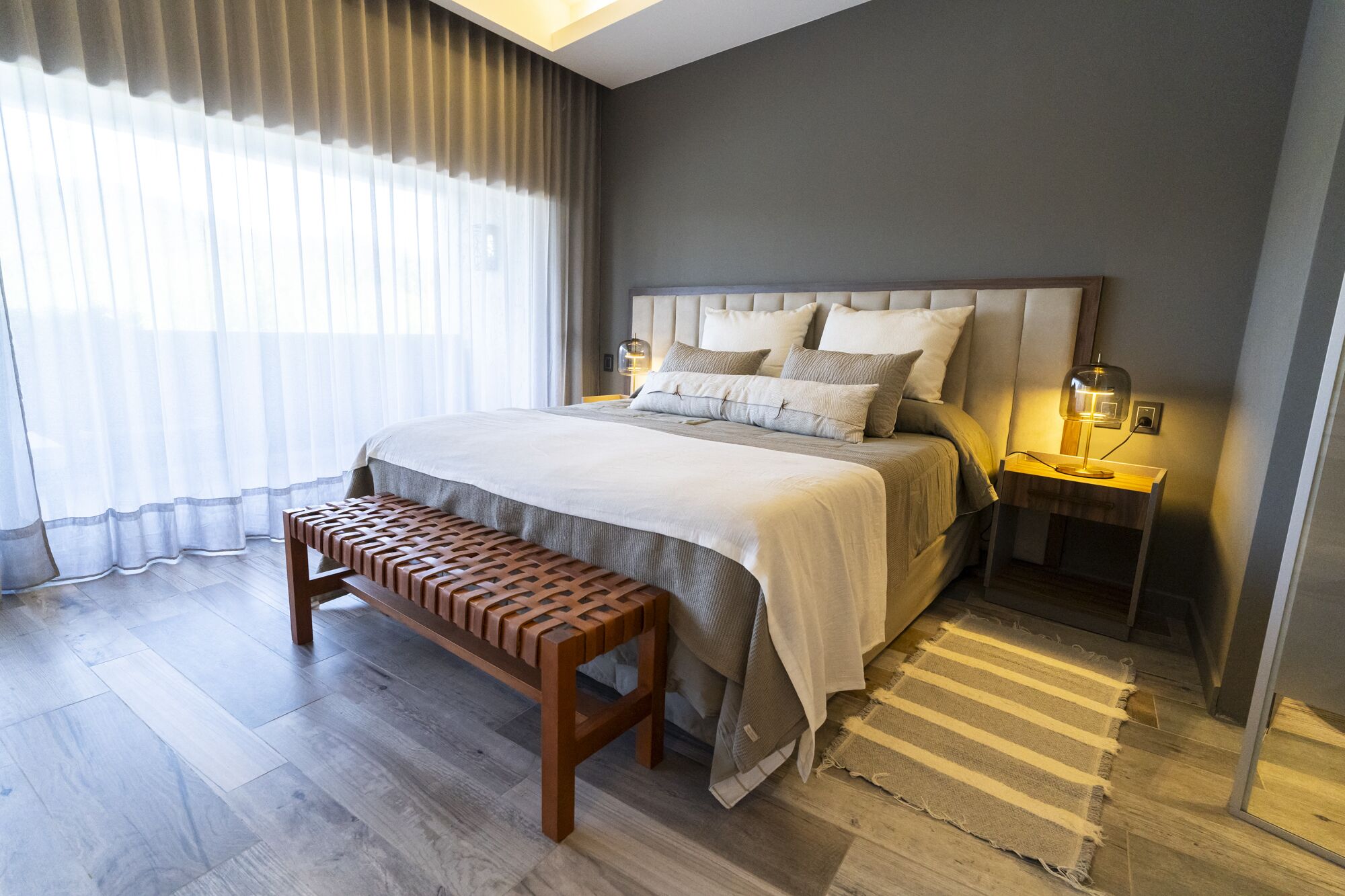 A modern bedroom with a large bed, wooden bench, two bedside tables with lamps, and sheer curtains.