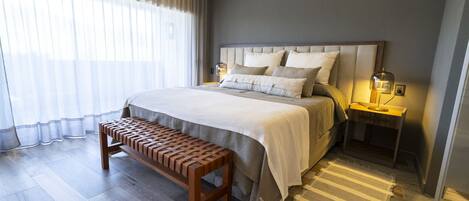 A modern bedroom with a large bed, wooden bench, two bedside tables with lamps, and sheer curtains.