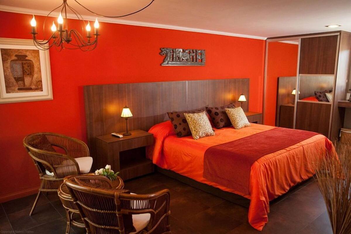 A hotel room with a red wall, a wooden headboard, a bed with orange bedding, two nightstands with lamps, wicker chairs, and a chandelier.