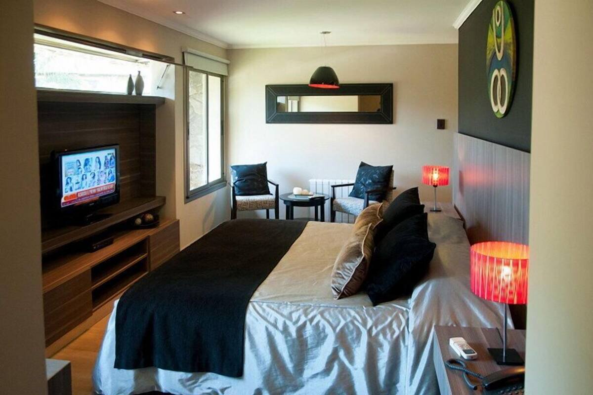 A modern hotel room with a large bed, a TV on a wooden stand, a small table with chairs, and a red lamp.