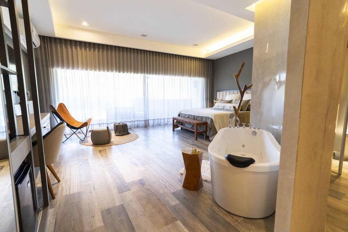 A modern hotel room with a large bed, a sitting area with a chair and ottoman, a large bathtub, and a view of the city through sheer curtains.