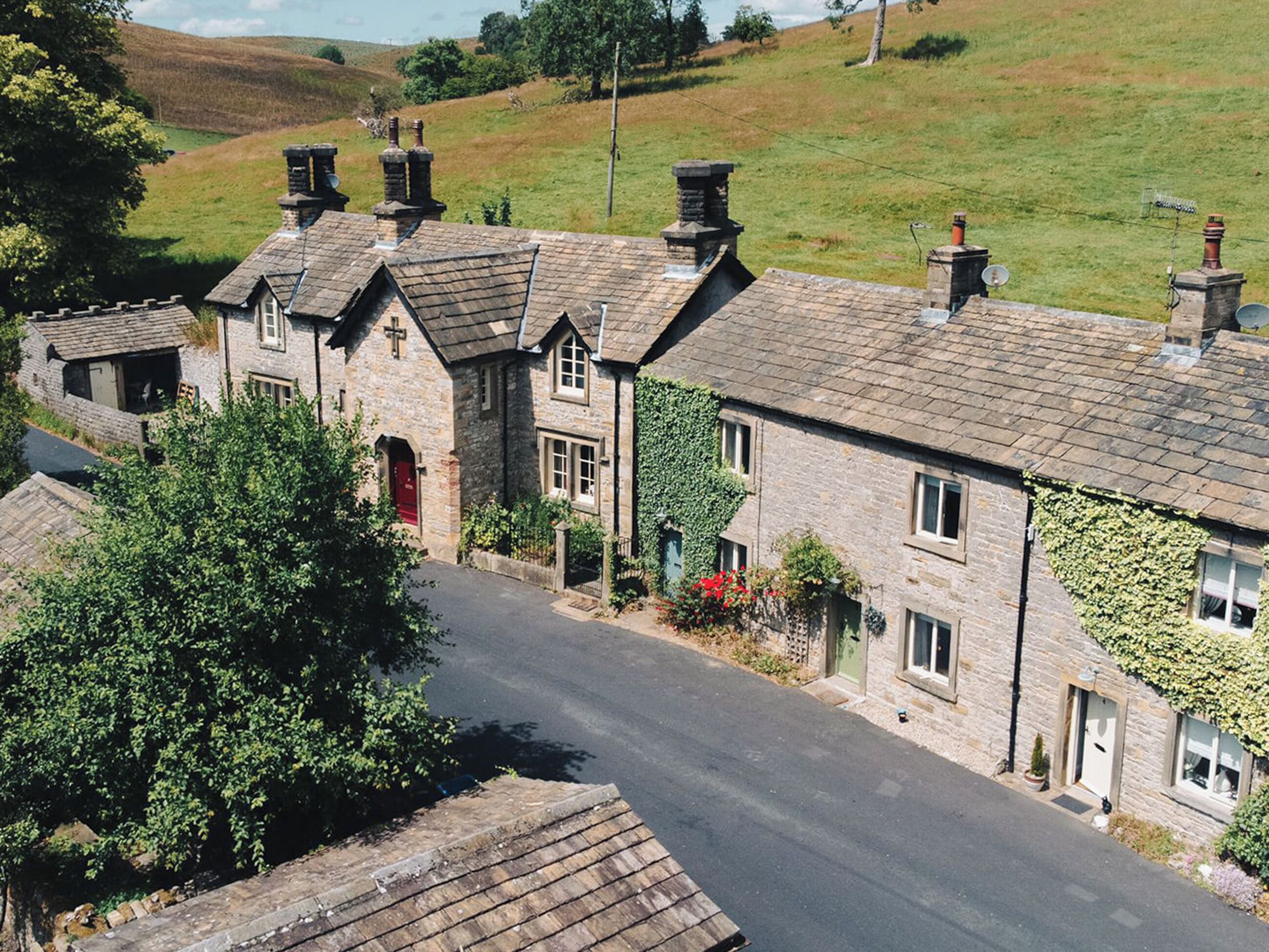 A row of stone houses with pitched roofs and chimneys, a narrow road, and a green hillside in the background.