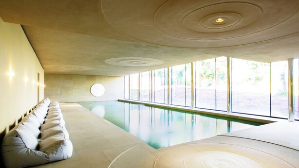 Piscina interior moderna com um longo banco ao longo de uma parede, janelas amplas e um design de teto único.