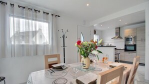 Un comedor con una mesa dispuesta para dos personas, un jarrón de flores y una vista de una cocina moderna al fondo.