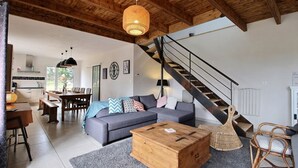 Un amplio salón con techo de madera, un sofá gris, una mesa de centro de madera, sillas de mimbre y una escalera.