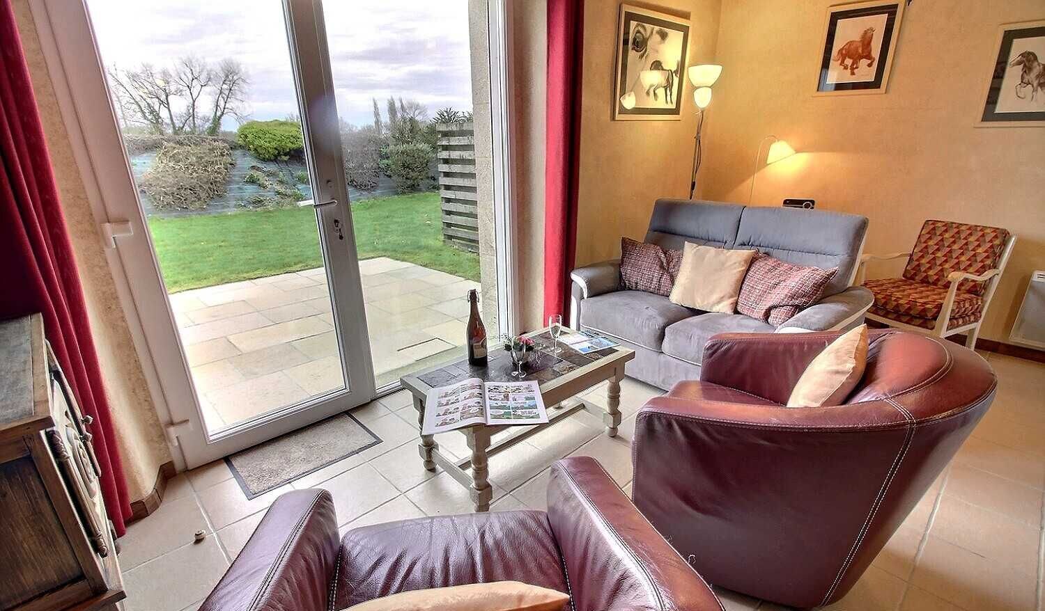 Un salon comprenant un canapé, un fauteuil et une table avec un magazine. Une porte vitrée donne accès à un jardin.