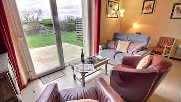 A living room with a sofa, armchair, and a table with a magazine. There is a glass door leading to a garden.
