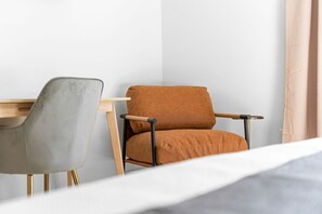 A corner of a room with a bed, a grey chair, and a brown chair with wooden legs.