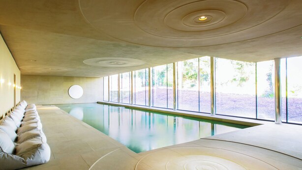Una piscina interior moderna con una amplia zona de asientos tipo banco, ventanas grandes y un diseño de techo original.