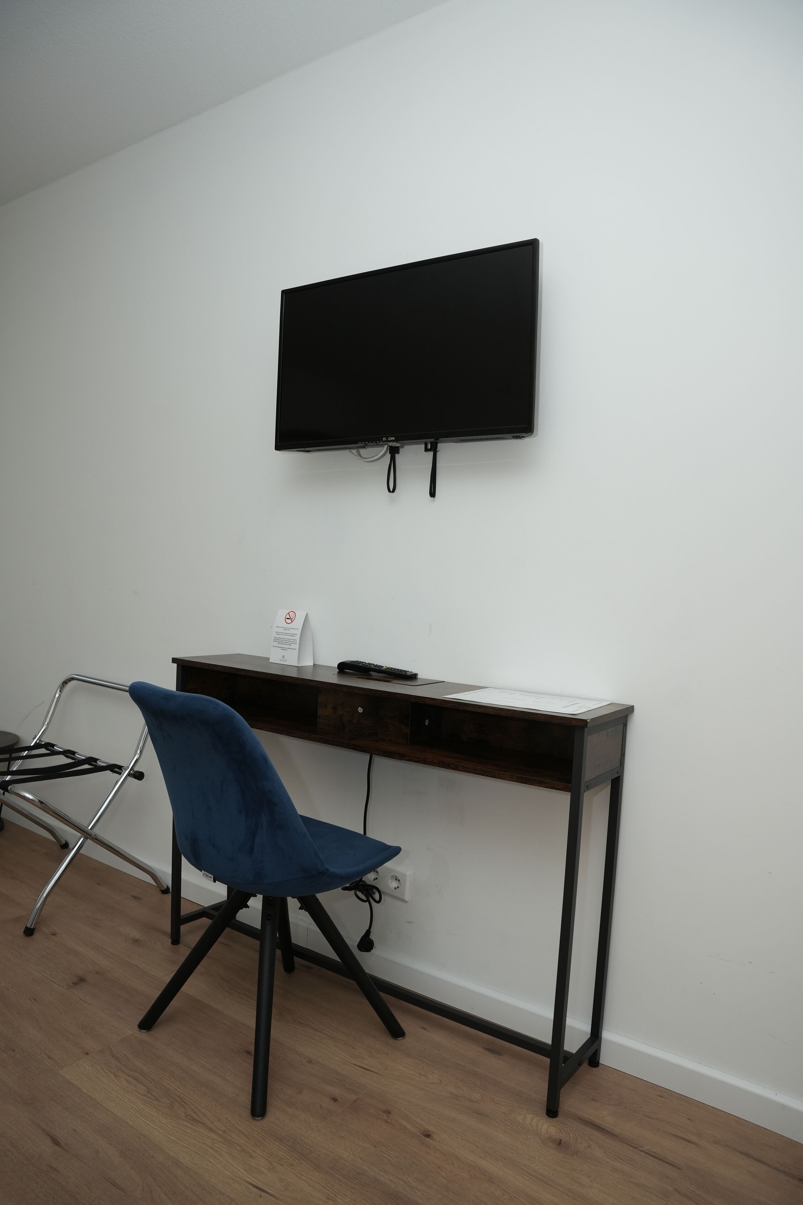 Una habitación con un escritorio de madera oscura, una silla azul, un televisor de pantalla plana montado en la pared y una pequeña señal en el escritorio.