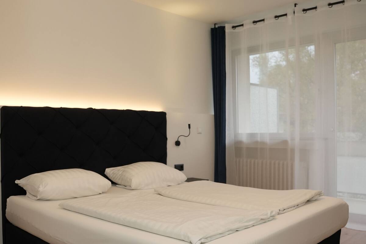 A bedroom with a bed, wooden flooring, a ceiling light, and a window with curtains.