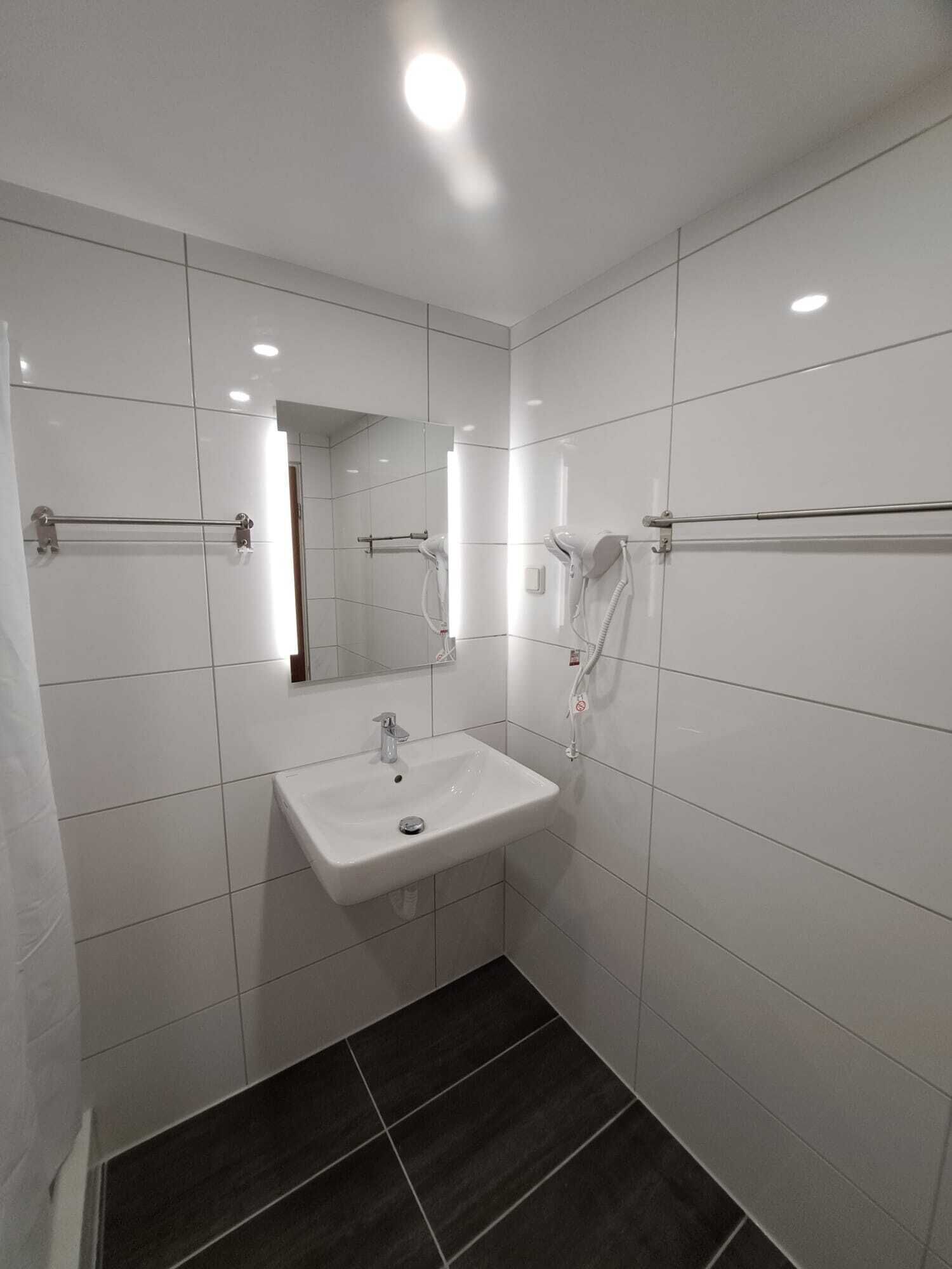 Un baño moderno con azulejos blancos, un lavamanos, un espejo y dos secadores de pelo fijados en la pared.