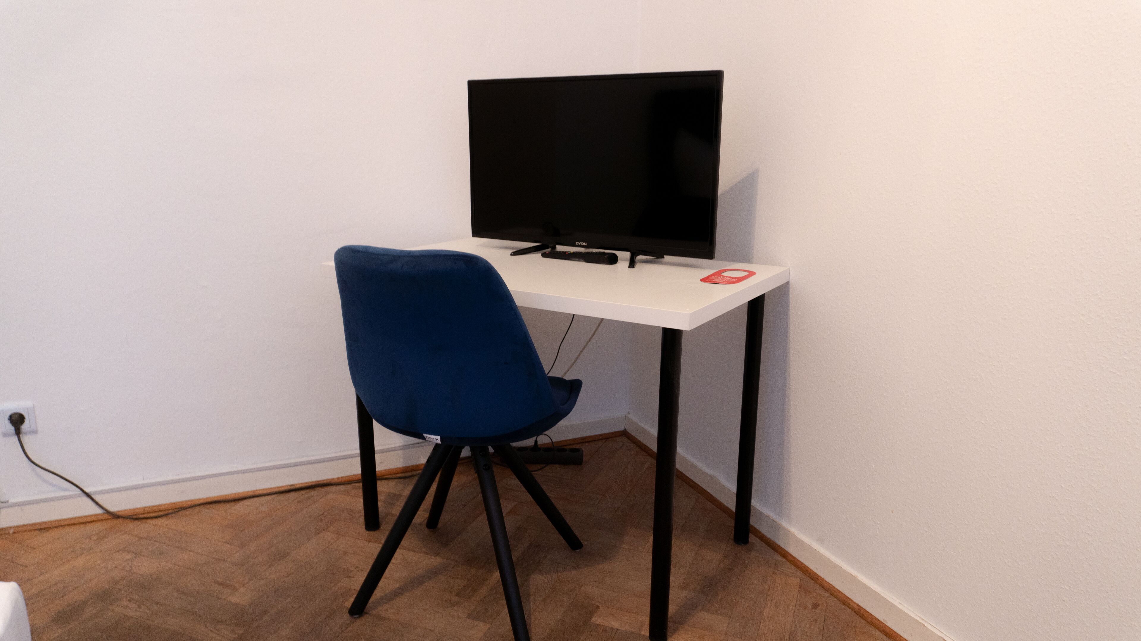 Un pequeño espacio de trabajo con un escritorio, una silla azul, un televisor negro y un objeto rojo sobre el escritorio.