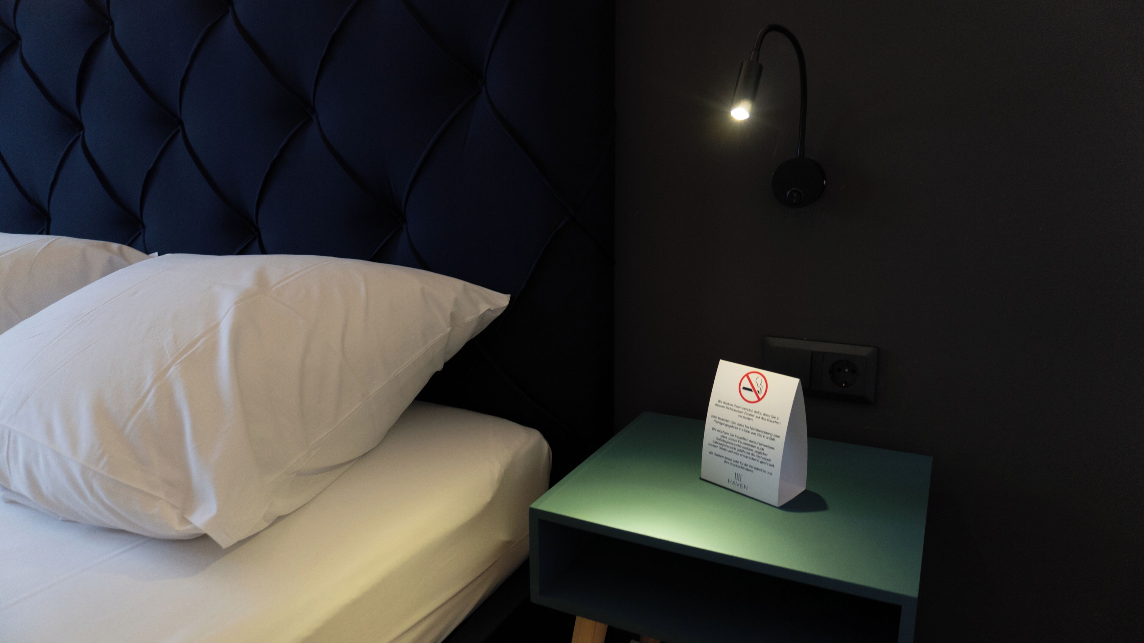 Un lit avec des oreillers blancs, une tête de lit sombre, une table de chevet avec un plateau vert et un panneau indiquant qu’il est interdit de fumer.