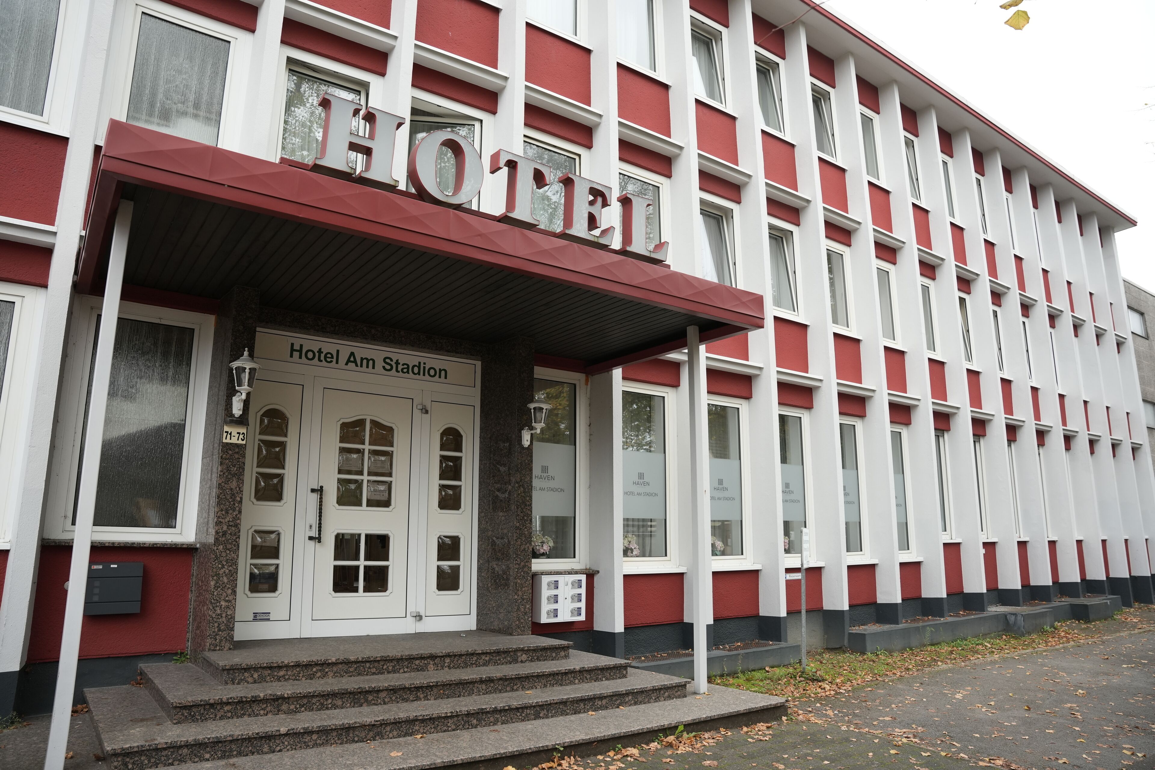 A hotel with a red and white facade, a prominent entrance with steps, and a sign reading "Hotel Am Stadion."