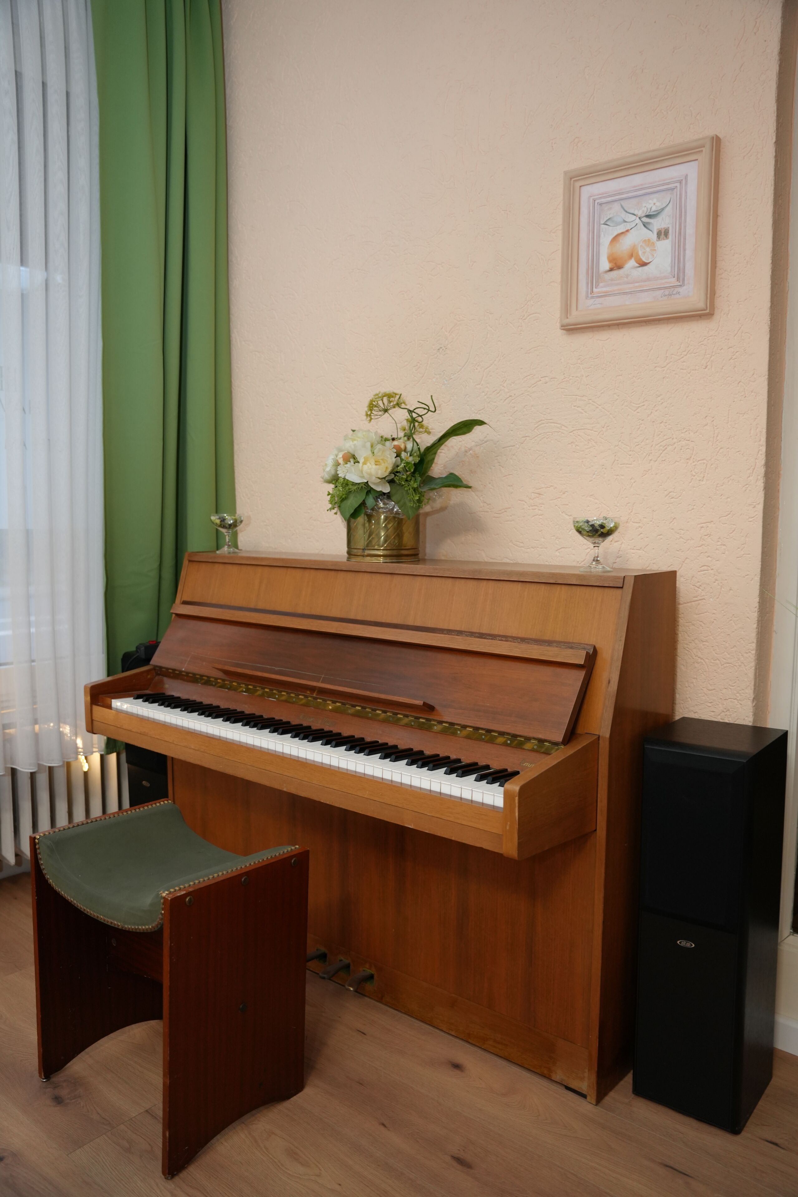 A wooden piano with a bench, a black speaker, a vase with flowers, and a framed picture on the wall.