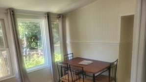 Une pièce avec une table en bois et quatre chaises, une fenêtre avec des rideaux et une vue sur la verdure à l’extérieur.