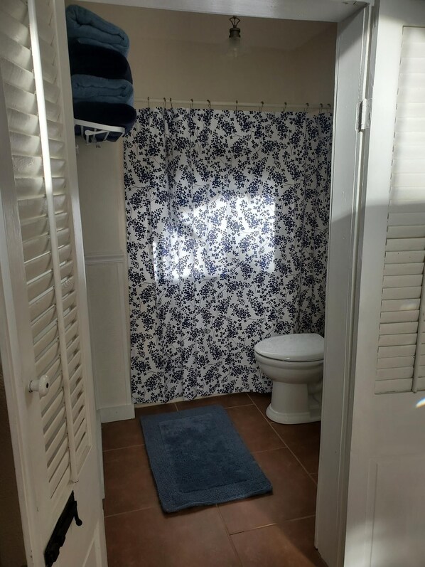 A bathroom with a toilet, a shower curtain with a floral pattern, a blue bath mat, and a white door with a towel rack above it. - Claiborne Suite in Old Mandeville (Mandeville)