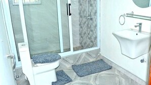 A bathroom with a glass shower enclosure, a toilet, a sink, and a mirror. - Tamarind Guesthouse (Aburi)
