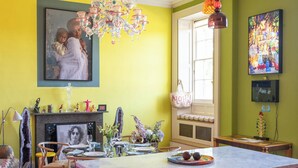 Une pièce lumineuse aux murs jaune vif, dotée d’une cheminée, d’un coin repas et d’une cuisine avec un meuble de couleur rose.
