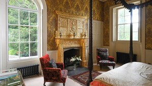 Une chambre dotée d’un lit à baldaquin, d’une cheminée, de deux fauteuils et d’une grande fenêtre.