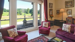 Une pièce comprenant deux fauteuils rouges, un canapé, une grande fenêtre donnant sur un paysage verdoyant et un lustre.