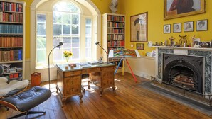 Une pièce avec une cheminée, des étagères à livres, un bureau avec une lampe et une grande fenêtre.