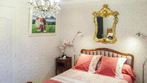Un dormitorio con cama de madera, papel tapiz floral, un candelabro, un espejo y una mesita de noche con lámpara.