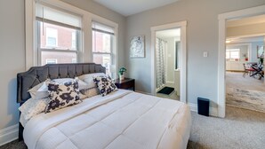 Quarto com cama grande, mesa de cabeceira e janela com persianas. Uma porta dá acesso a outro cômodo.