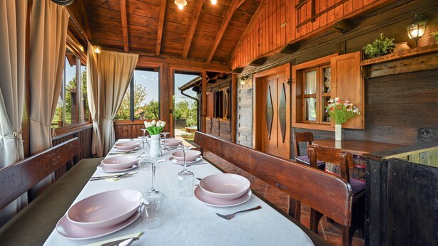 Une cabane en bois dotée d’un coin repas aménagé pour quatre personnes, avec une longue table ornée d’assiettes roses et de verres à vin, des bancs en bois et une vue sur l’extérieur.