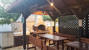 An outdoor patio with a stone floor, wooden tables and benches, a brick oven, and a sink area.
