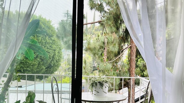 A balcony with a table and chair, surrounded by greenery and a view of trees and a building. - West Hollywood, Hollywood Hills, Sunset Strip House with Beautiful Views (Beverly Hills)