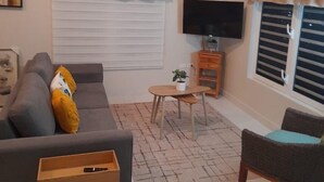 A living room with a grey sofa, a wooden side table, a small wooden table with a plant, a TV on a stand, and a window with blinds. - B’s Hidden Paradise (Mary’s Fancy)