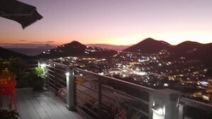 A balcony with a railing, potted plants, and a view of a cityscape at dusk. - B’s Hidden Paradise (Mary’s Fancy)