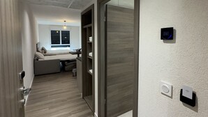 Een moderne hotelgang met een houten vloer, een bed op de achtergrond en een digitale thermostaat aan de muur.