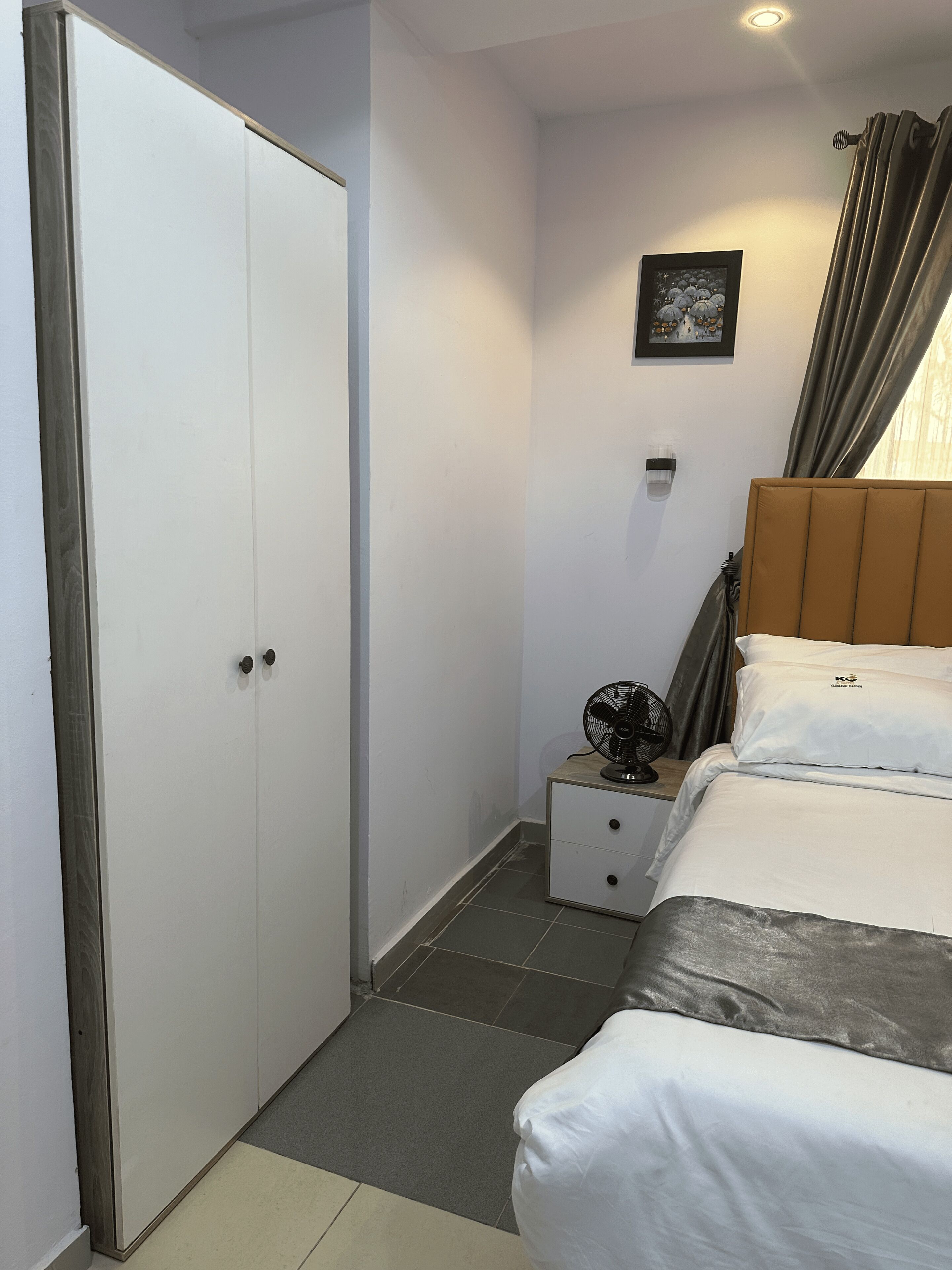 A bedroom with a bed, a bedside table with a fan, a nightstand, a wardrobe, and a picture on the wall.