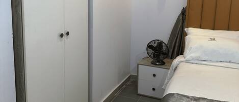 Un dormitorio con una cama, una mesita de noche con un ventilador, un armario y un cuadro en la pared.