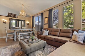 A living room with a brown leather sofa, a dining table with chairs, a chandelier, and large windows with curtains. - PRIVATE RESORT RETREAT - Pool, Hot Tub, Sauna & Games (Gilbert)